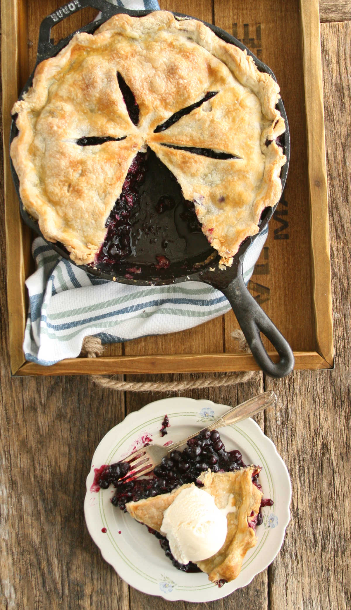 Blueberry pie in cast iron skillet, slice of pie on small white plate with fork.