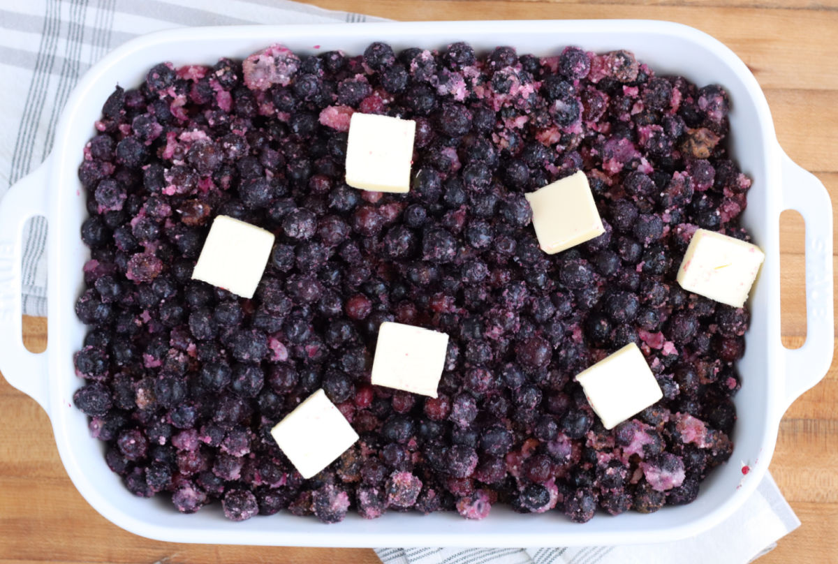 Blueberries in white rectangle baking dish topped with butter on wooden cutting board.