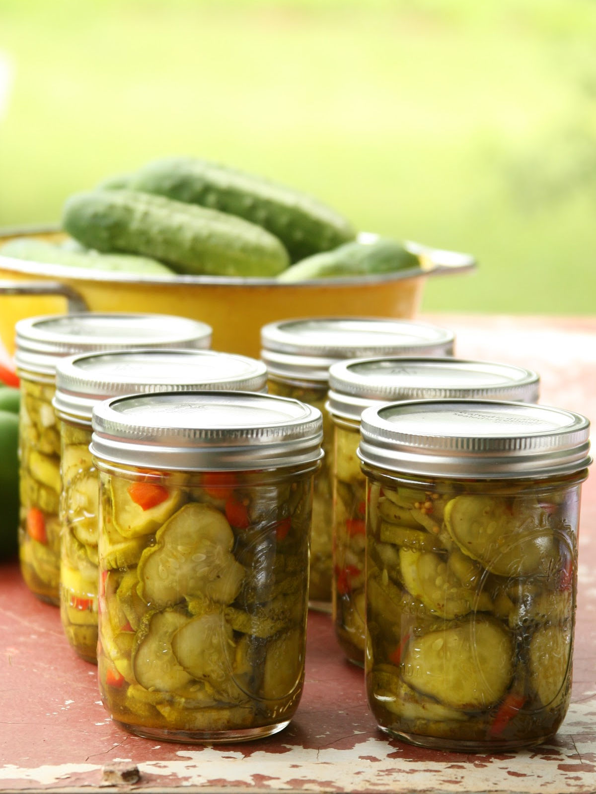 Pint jars of pickles on outdoor patio, yellow color strainer filled with cucumbers in background.