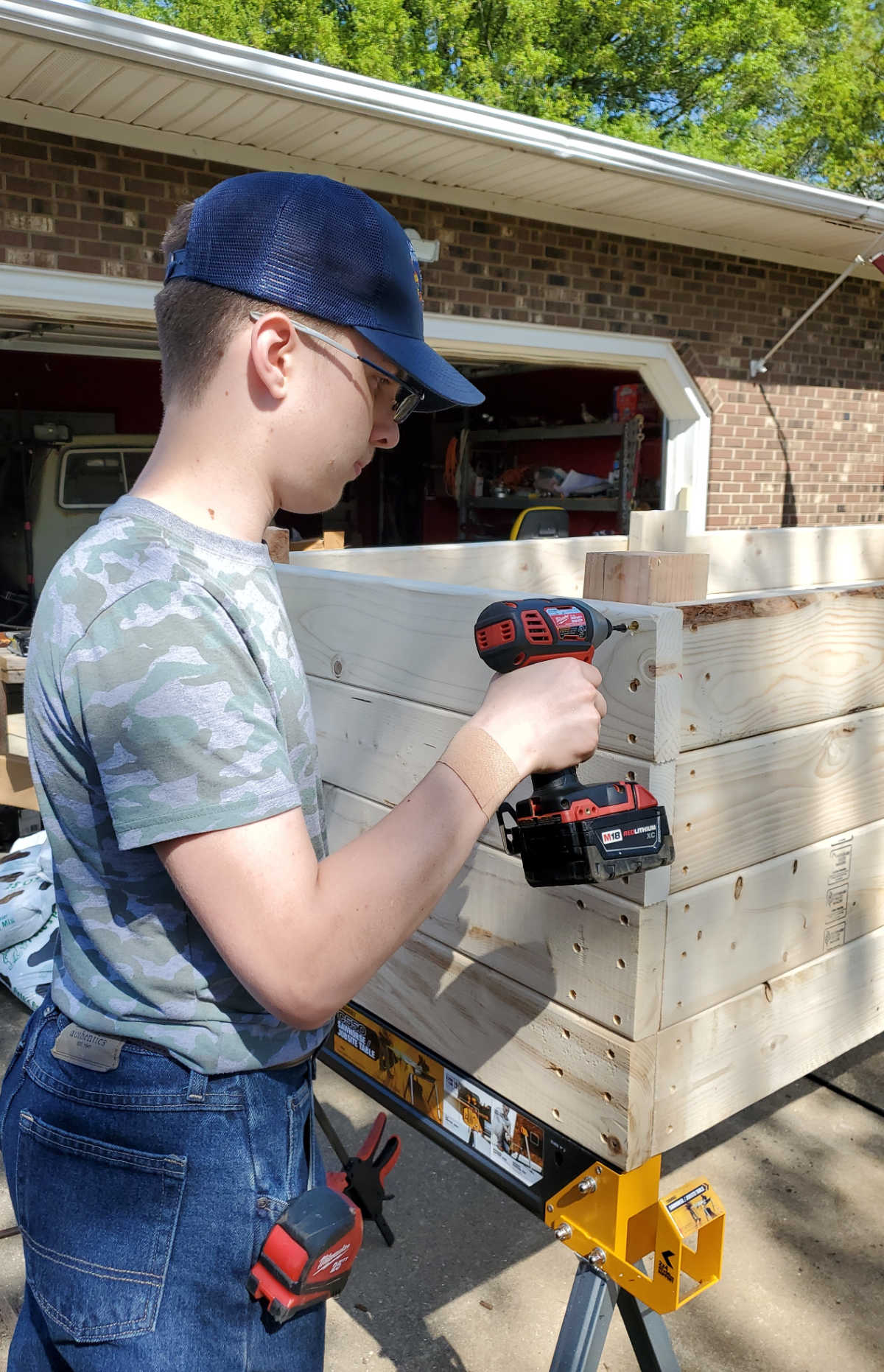 Young boy with jeans and blue baseball cap using drill to put screws into wood on DIY raised bed.