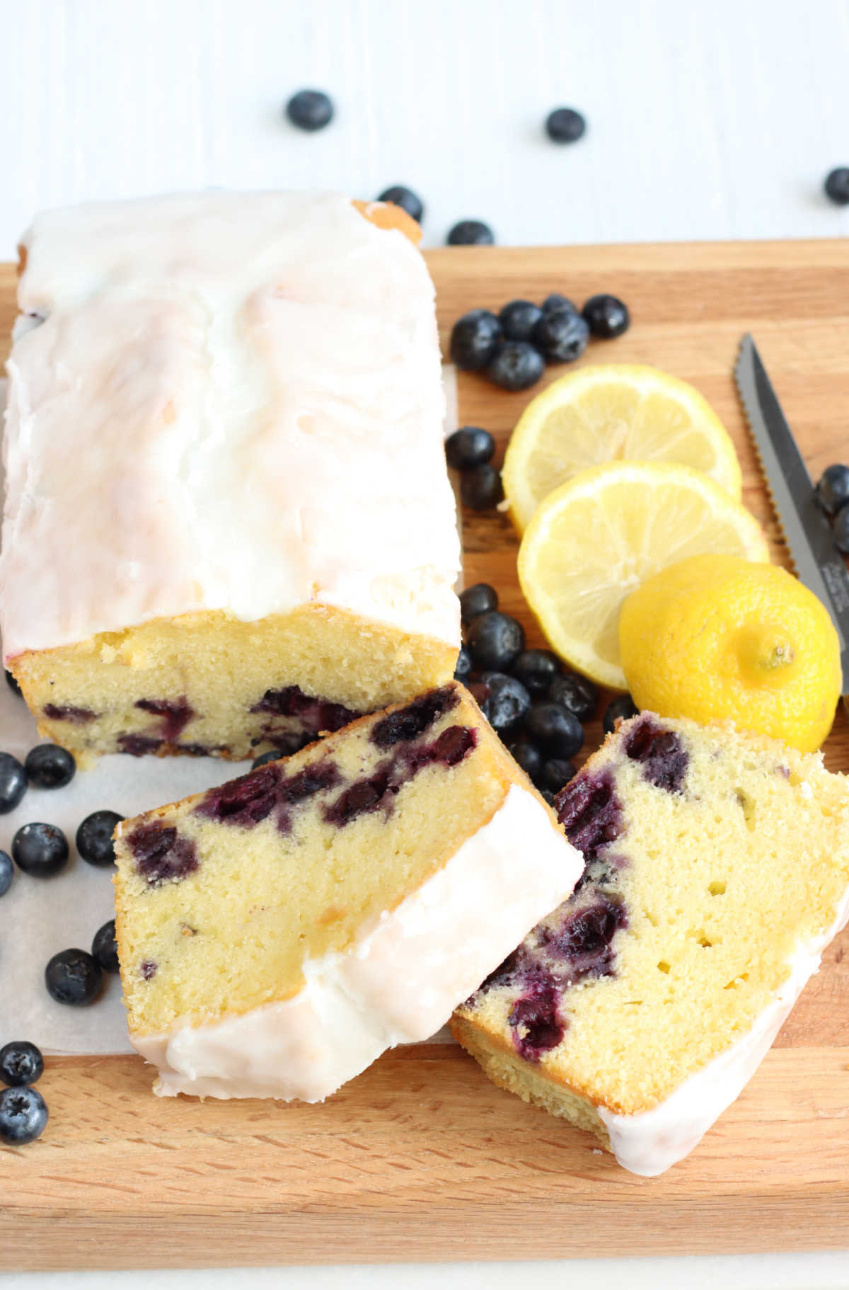 Lemon loaf cake with blueberries on wooden cutting board, lemon slices to right.