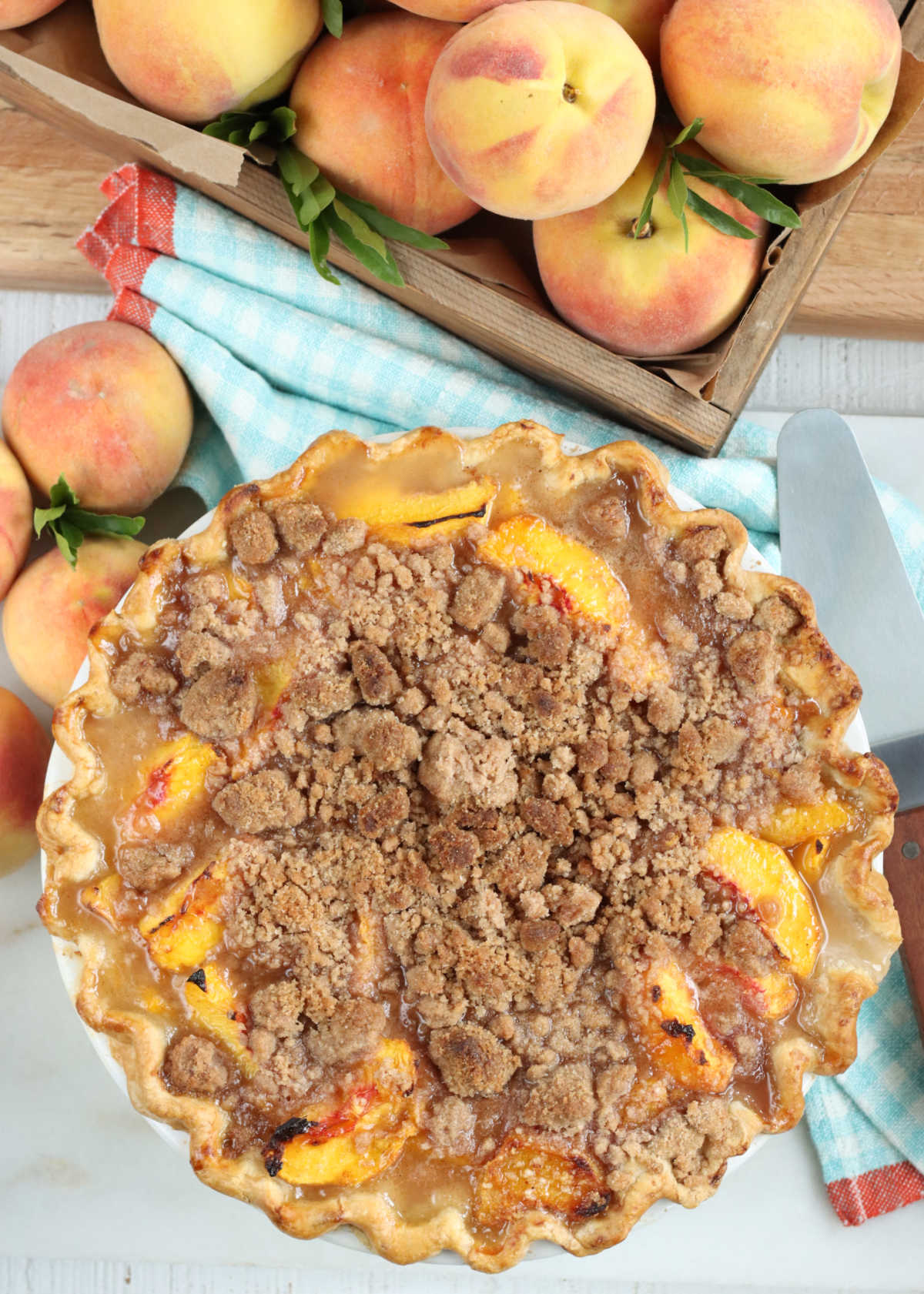 Crumb peach pie on white marble, fresh peaches in wooden box in background.