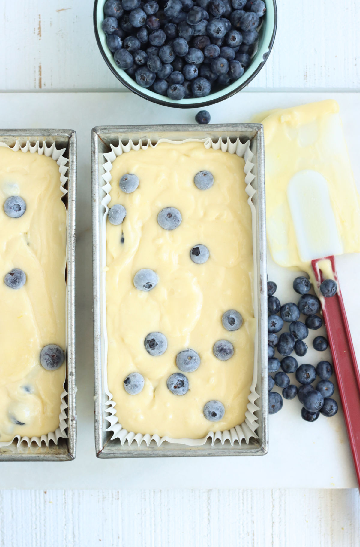 Two metal loaf pans with lemon blueberry bread batter, loose blueberries and red handle spatula to right.