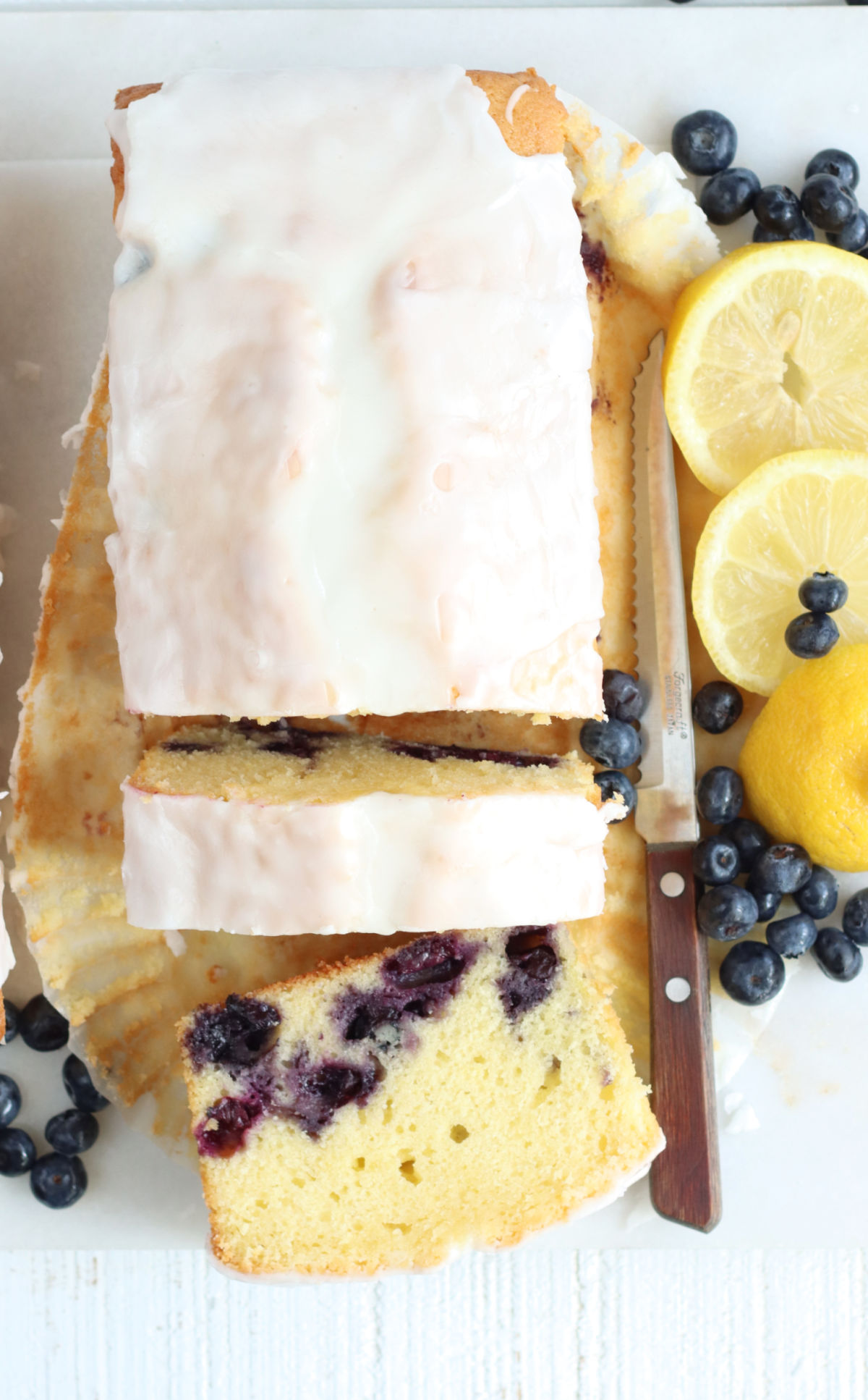 Partially sliced lemon blueberry loaf cake on white marble, slices of lemons, knife, fresh blueberries to right.