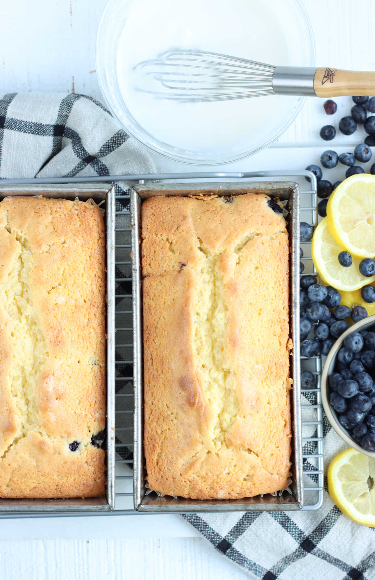 Two lemon loaf cakes in metal loaf pans cooling, lemon slices to right.