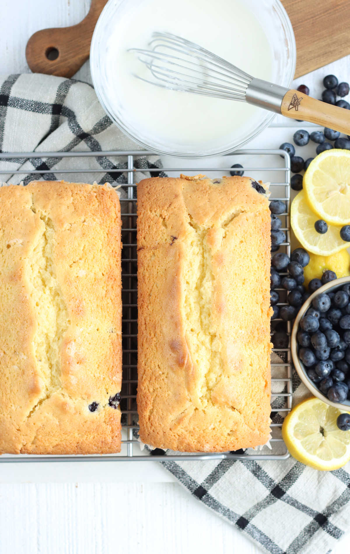 Two lemon blueberry breads on metal baking rack, bowl of blueberries and lemon slices to right.