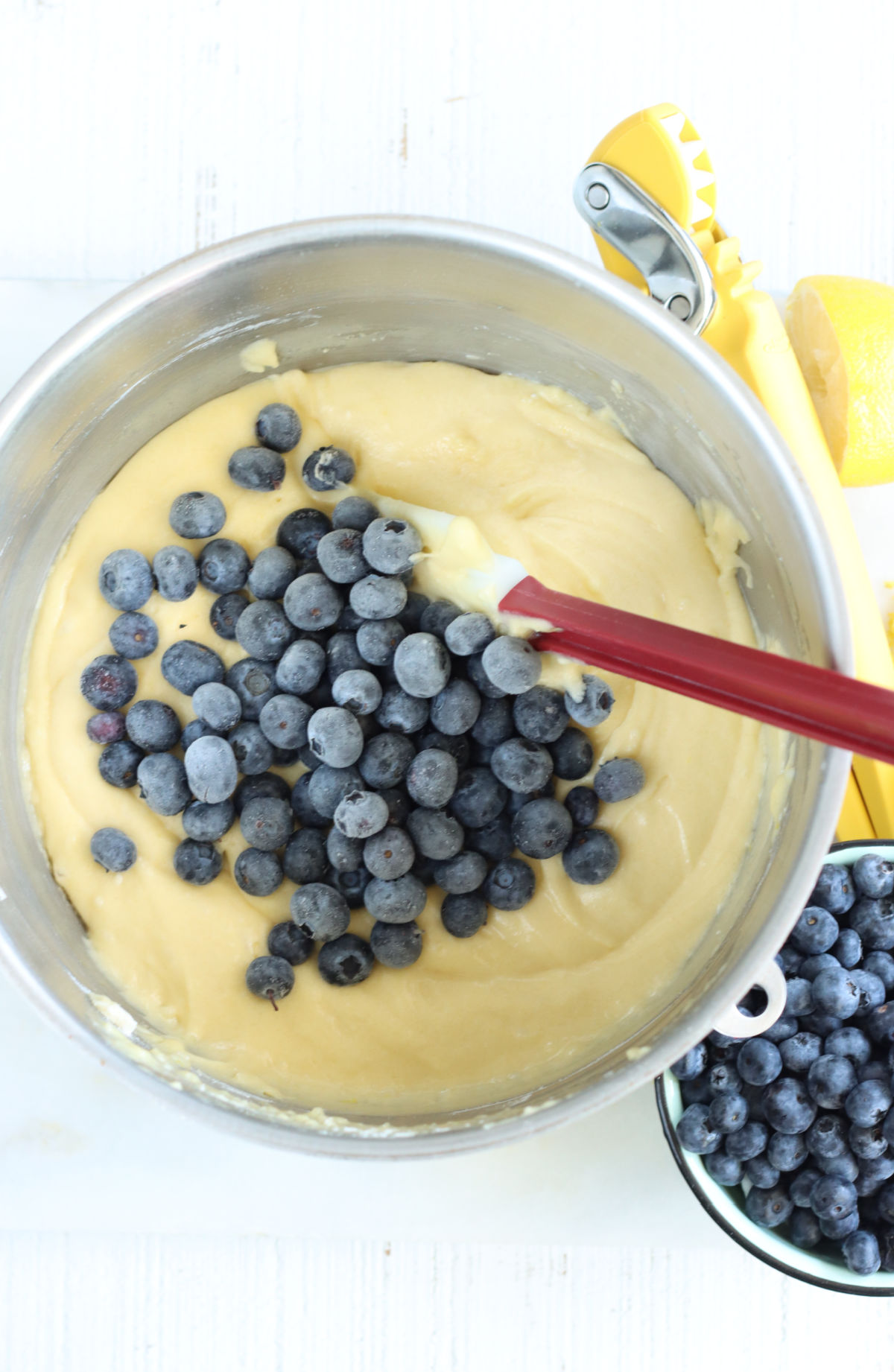 Lemon cake batter in metal mixing bowl with red handle spatula in bowl, fresh blueberries in small bowl to right.