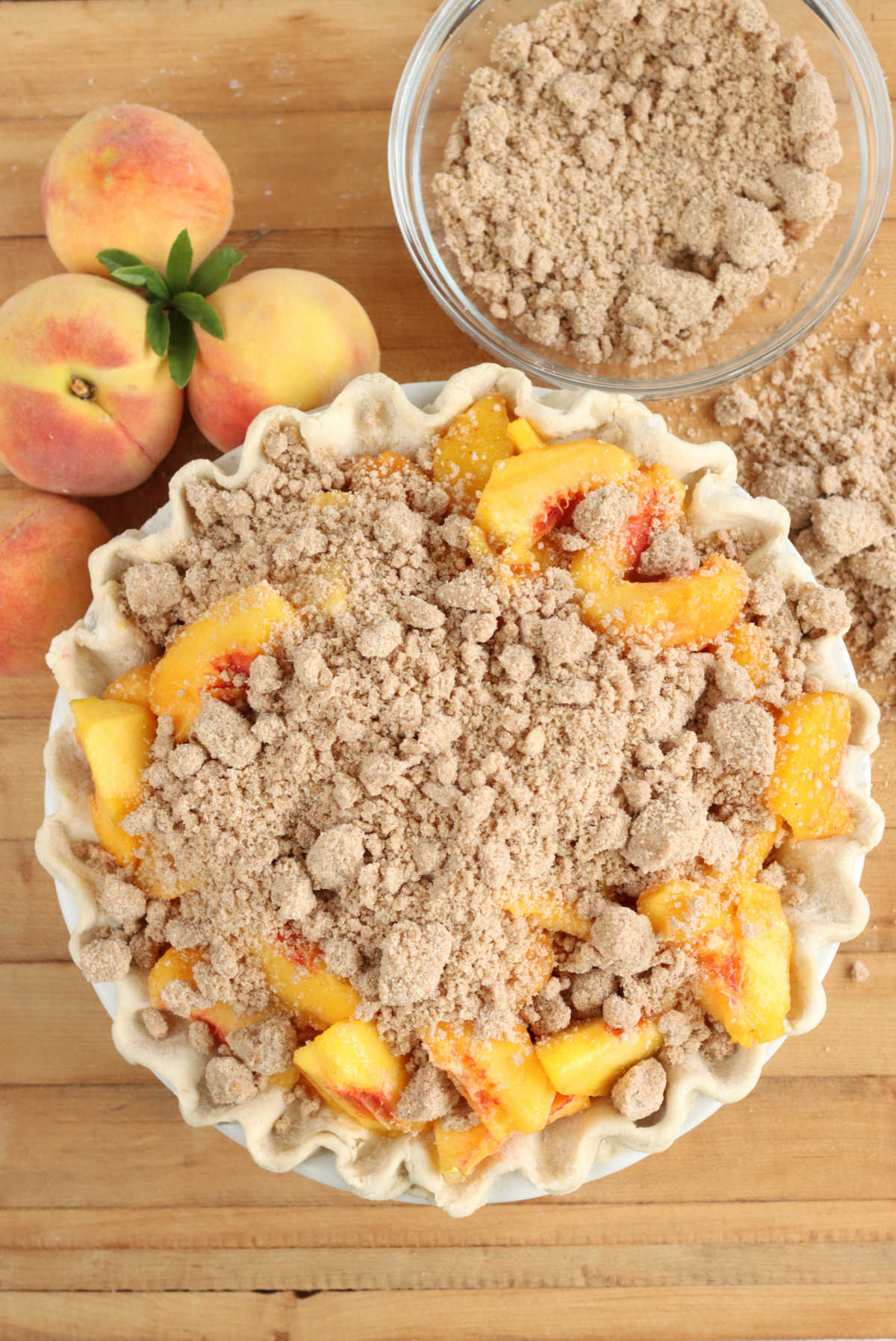 Unbaked peach pie with crumb topping on butcher block, fresh peaches around, clear glass bowl of crumb topping.