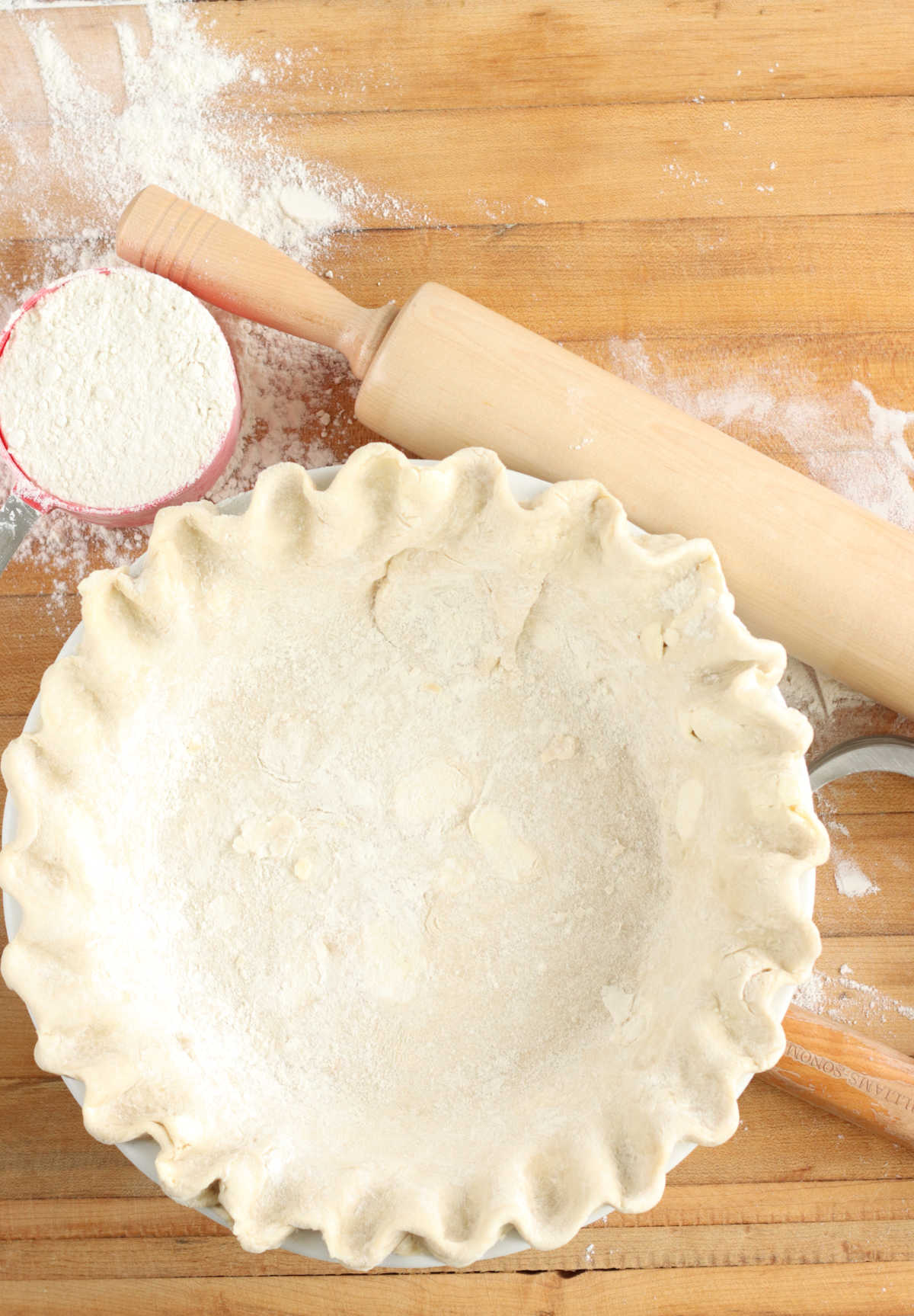 Unbaked pie shell on wooden cutting board with wooden rolling pin and cup of flour.