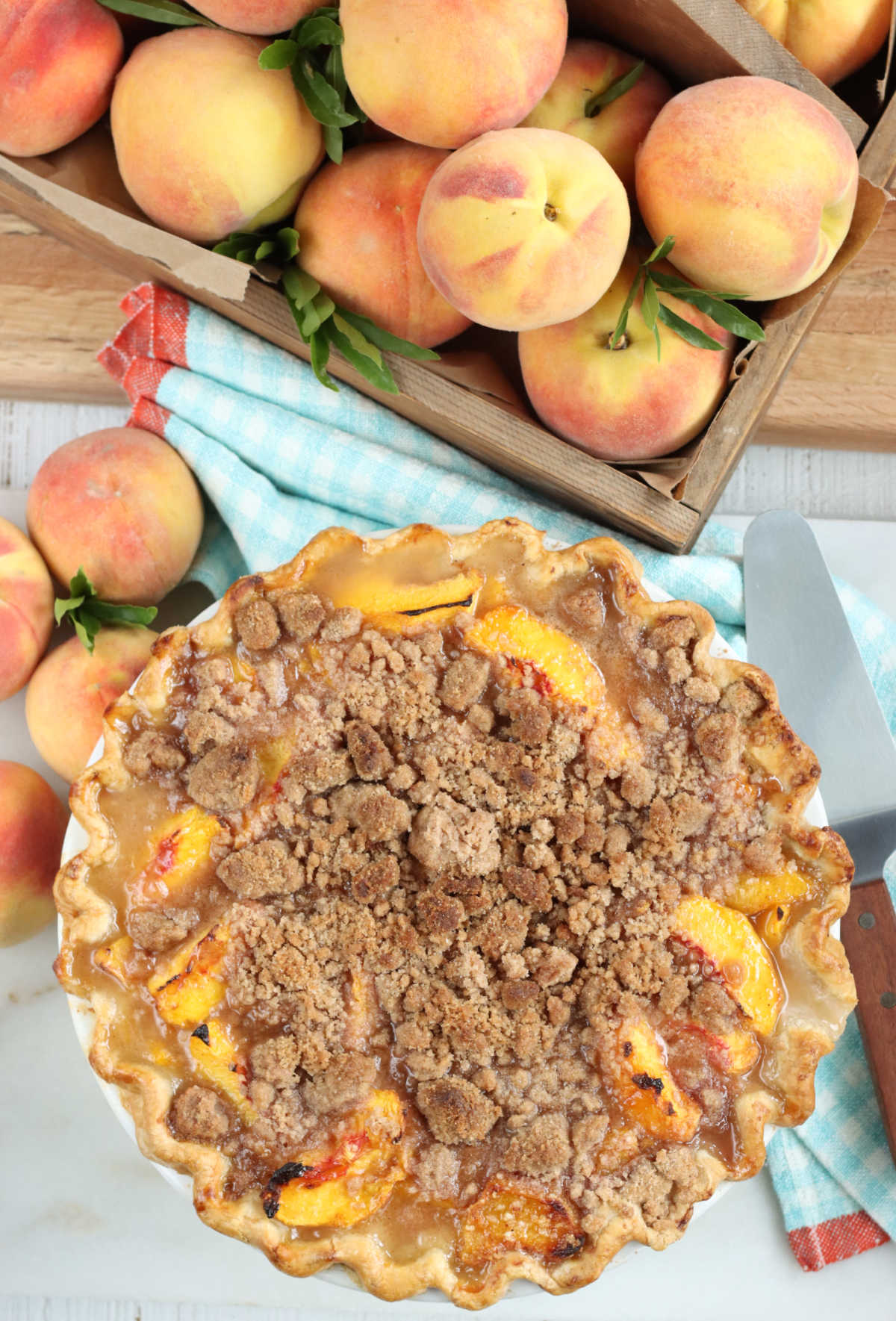 Peach pie with crumb topping on white marble, peaches in wooden box.