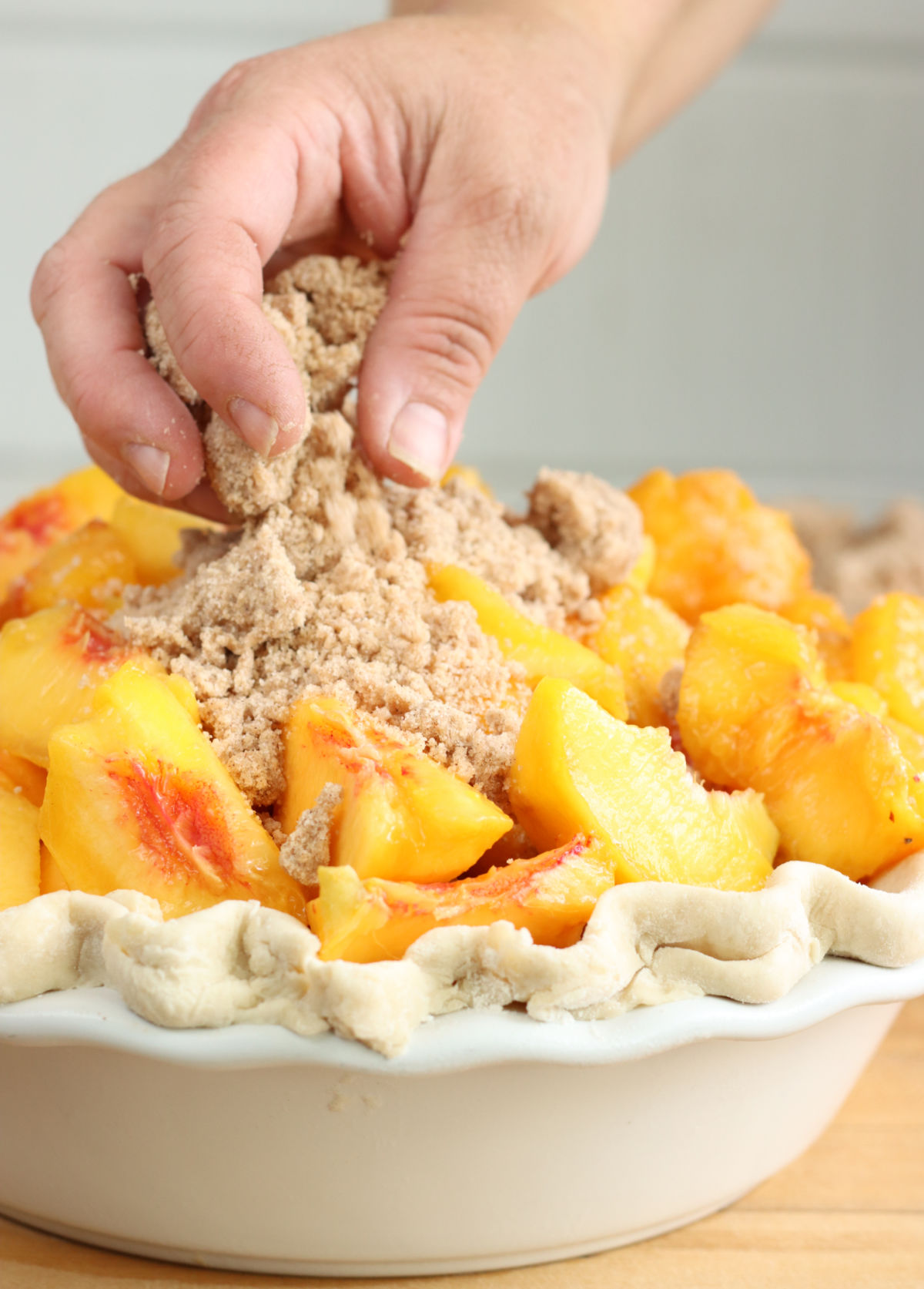 Hand putting crumble topping on peach pie of unbaked pie.