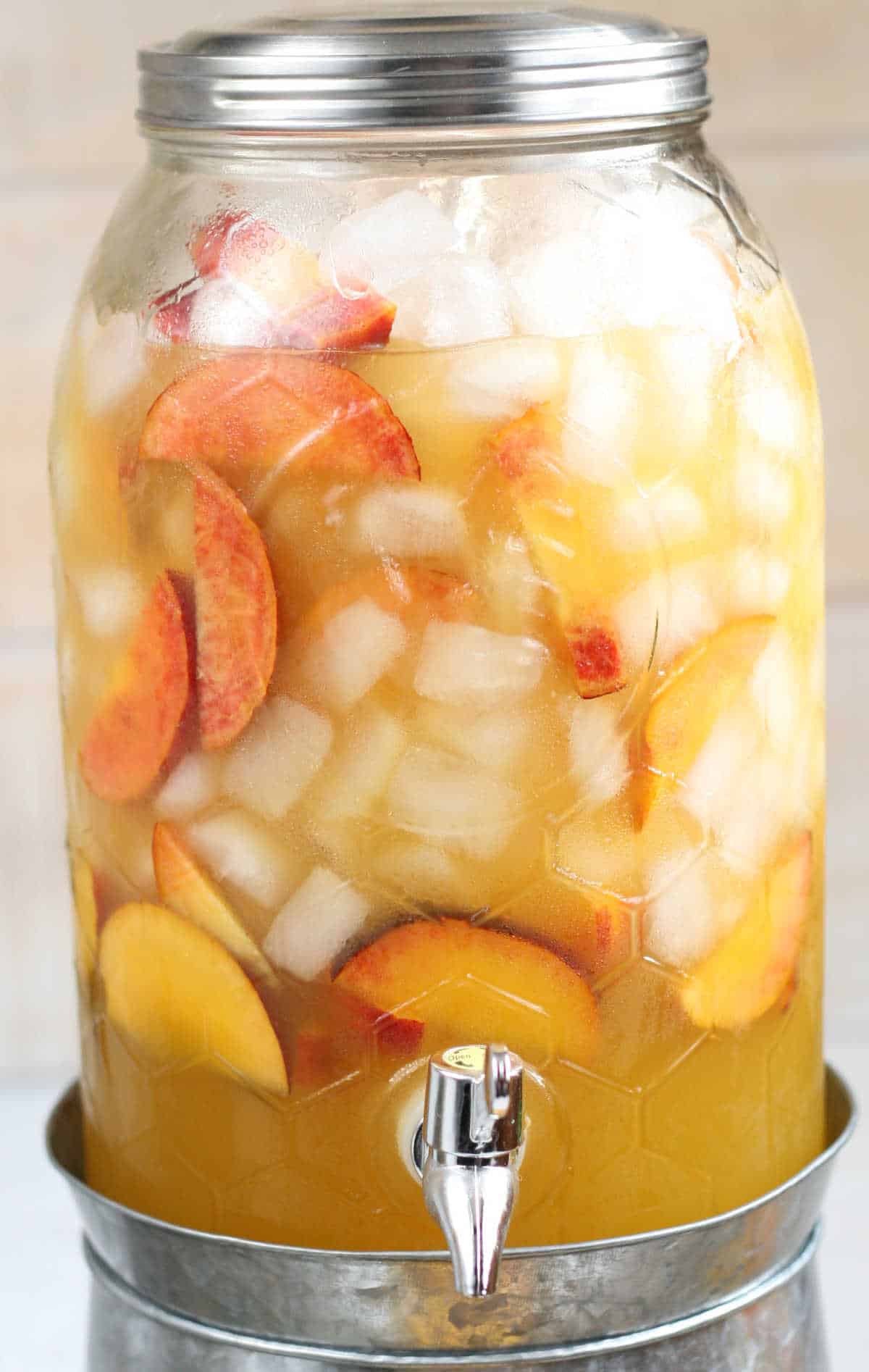 Close up of glass drink dispenser filled with white wine sangria, ice cubes, and peach slices.