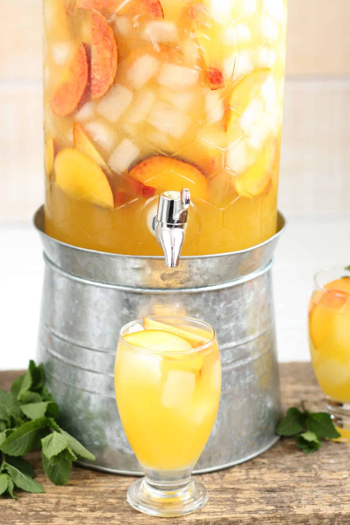 Summer sangria in clear glass drink dispenser, footed glass with ice cubes and slices of peaches in front.
