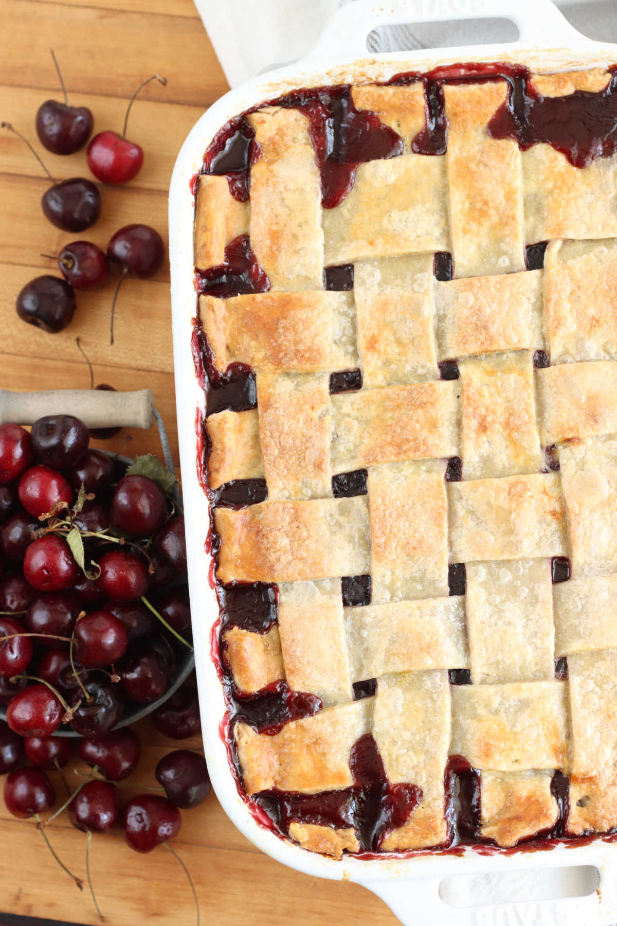 Cherry cobbler with lattice pie crust in white rectangle baking dish on wooden cutting board, fresh cherries to left.