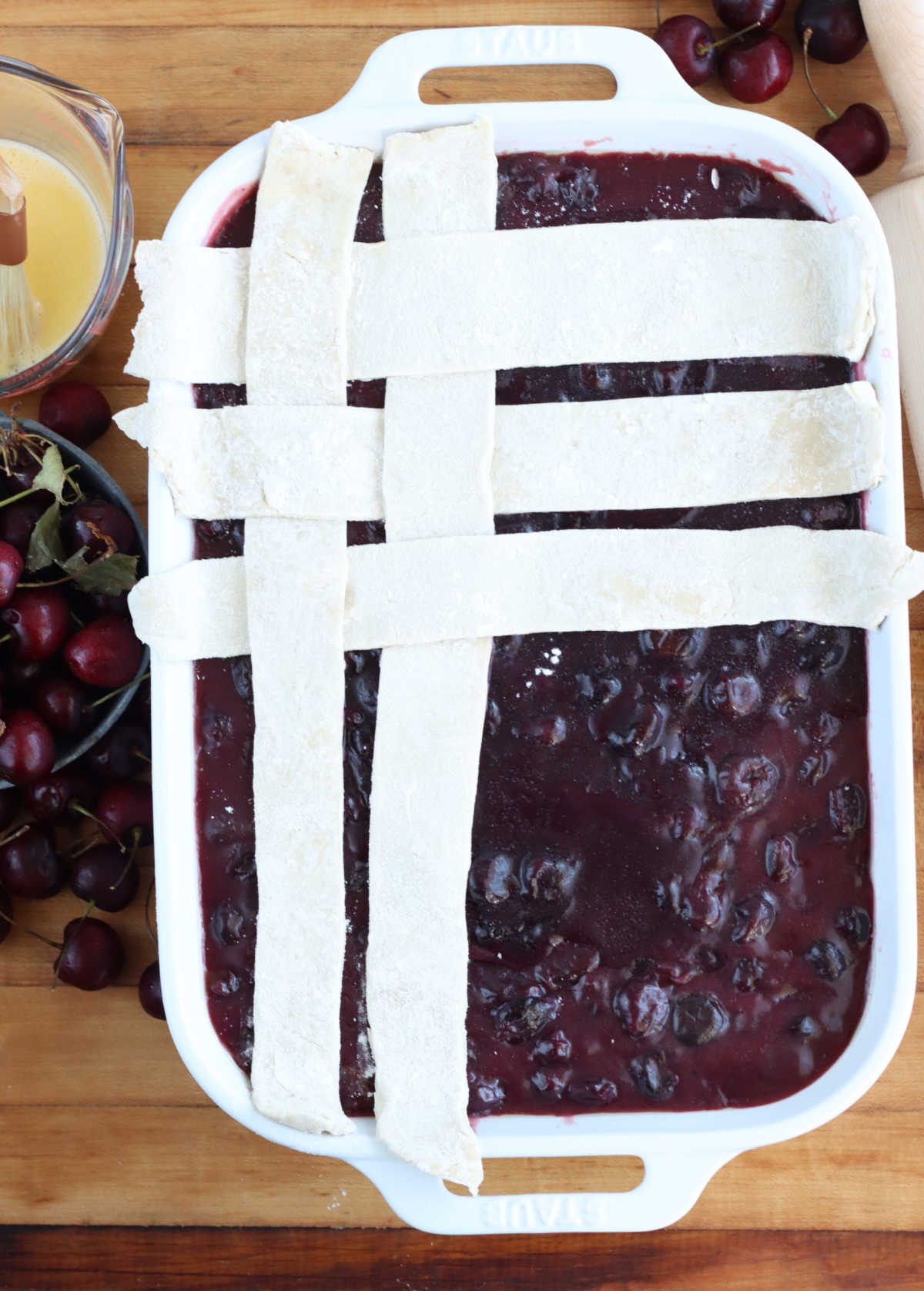 Weaving lattice pie crust on cherry cobbler in white rectangle baking dish.