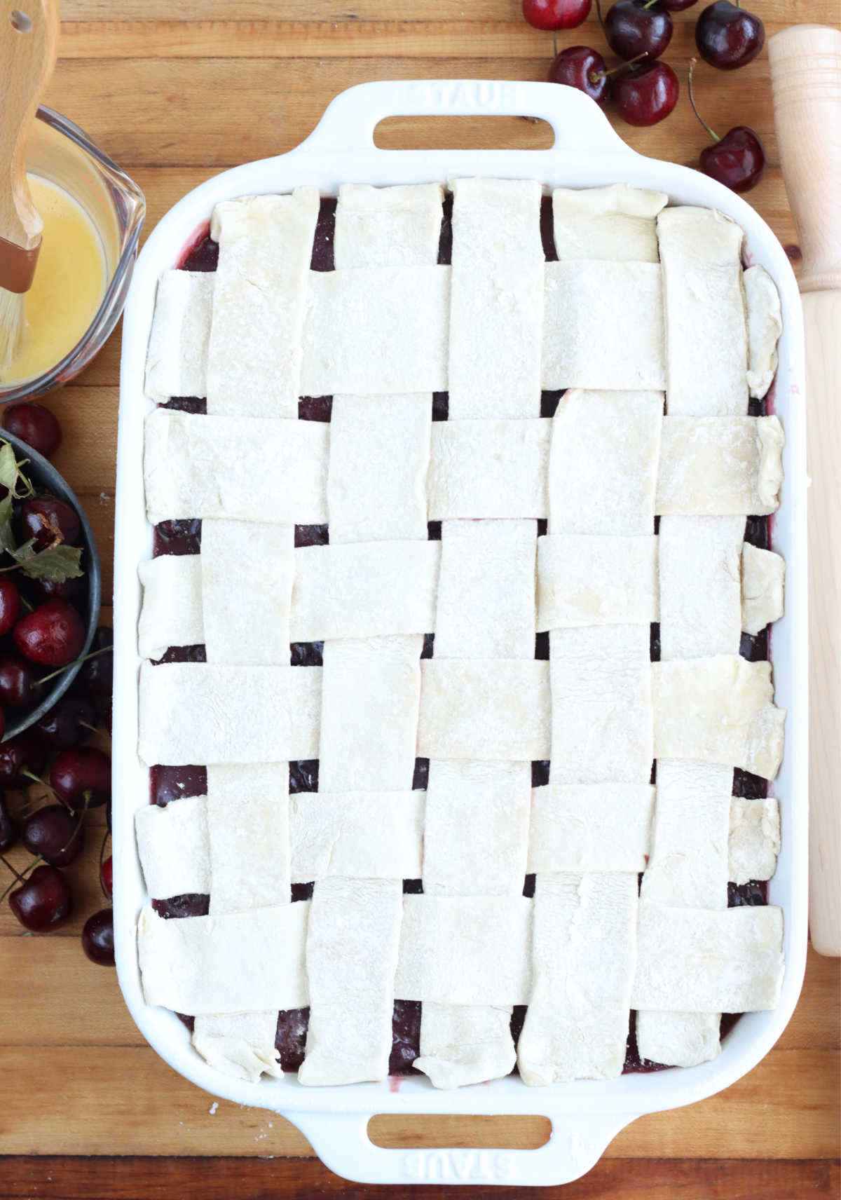 Cobbler with lattice pie crust in white rectangle baking dish, fresh cherries and egg wash to left.