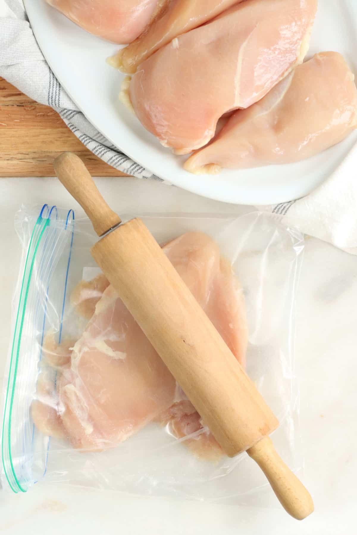 Chicken breast flattened in zip-style bag, small wooden rolling pin on white marble.