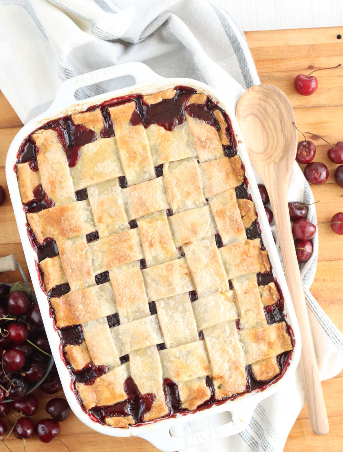 Cobbler with pie crust lattice weaved in white rectangle baking dish on wooden cutting board, fresh cherries to left, wooden spoon on right.
