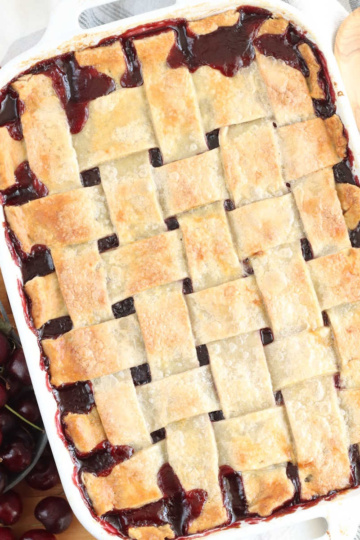 Cherry cobbler with lattice crust, fresh cherries around, wooden spoon.