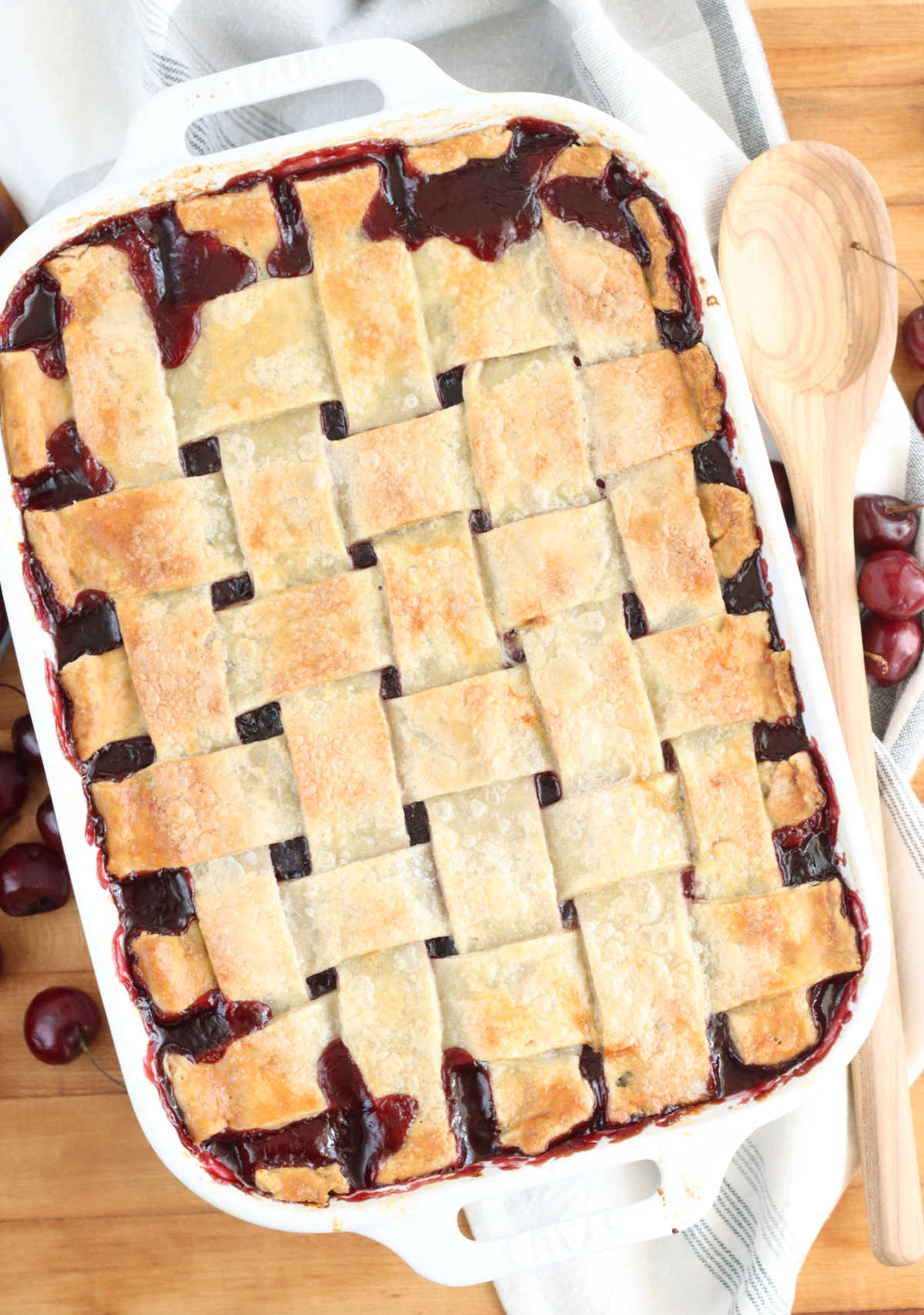 Cherry cobbler lattice pie crust in white rectangle baking dish on wooden cutting board, fresh cherries around.