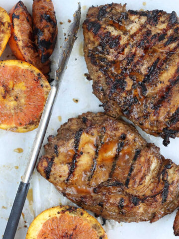 Two grilled chicken breasts on sheet pan with grilled orange slices and tongs.