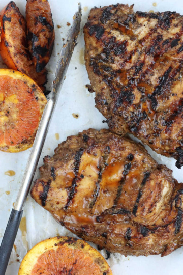 Two grilled chicken breasts on sheet pan with grilled orange slices and tongs.