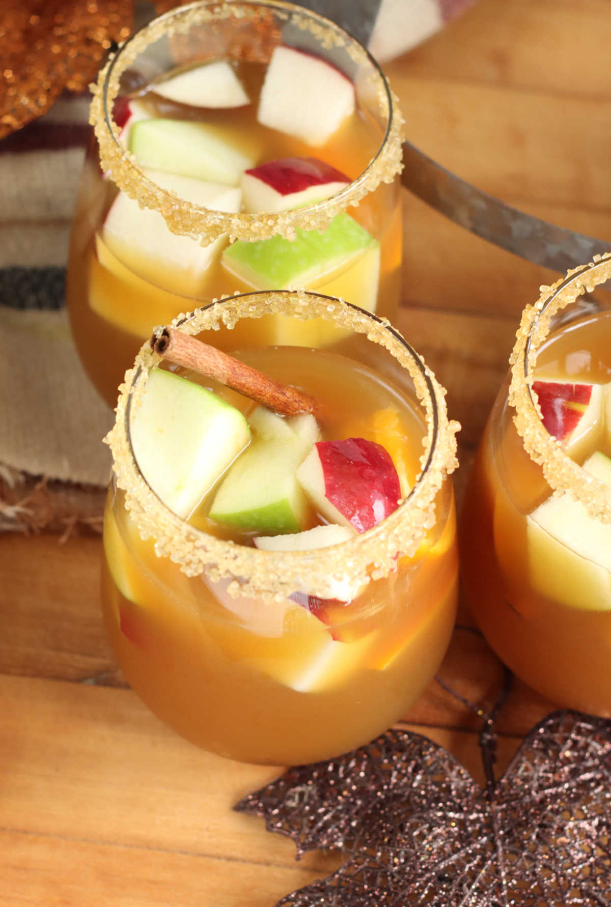 Stemless wine glasses with cider sangria, red and green apple chunks, oranges, cinnamon sticks, rims with raw sugar.