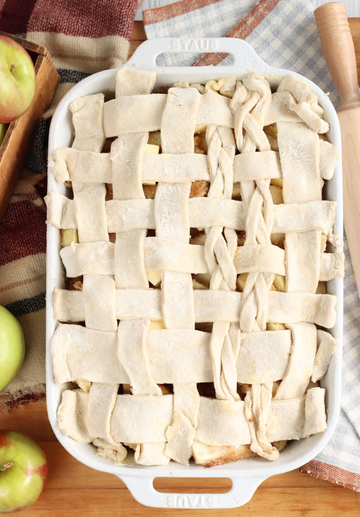 Unbaked apple cobbler with lattice pie crust in white baking dish on wooden cutting board, apples around.