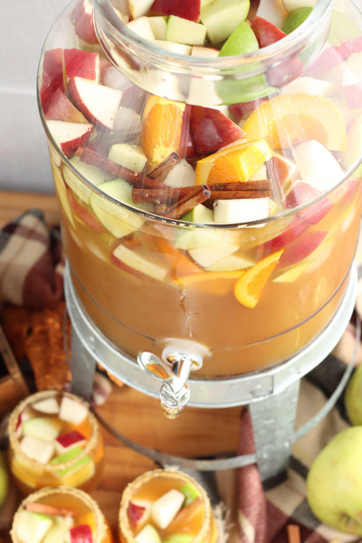 Clear drink dispenser with apple sangria, chunks of red, green apples, oranges, cinnamon sticks, stemless wine glasses below.
