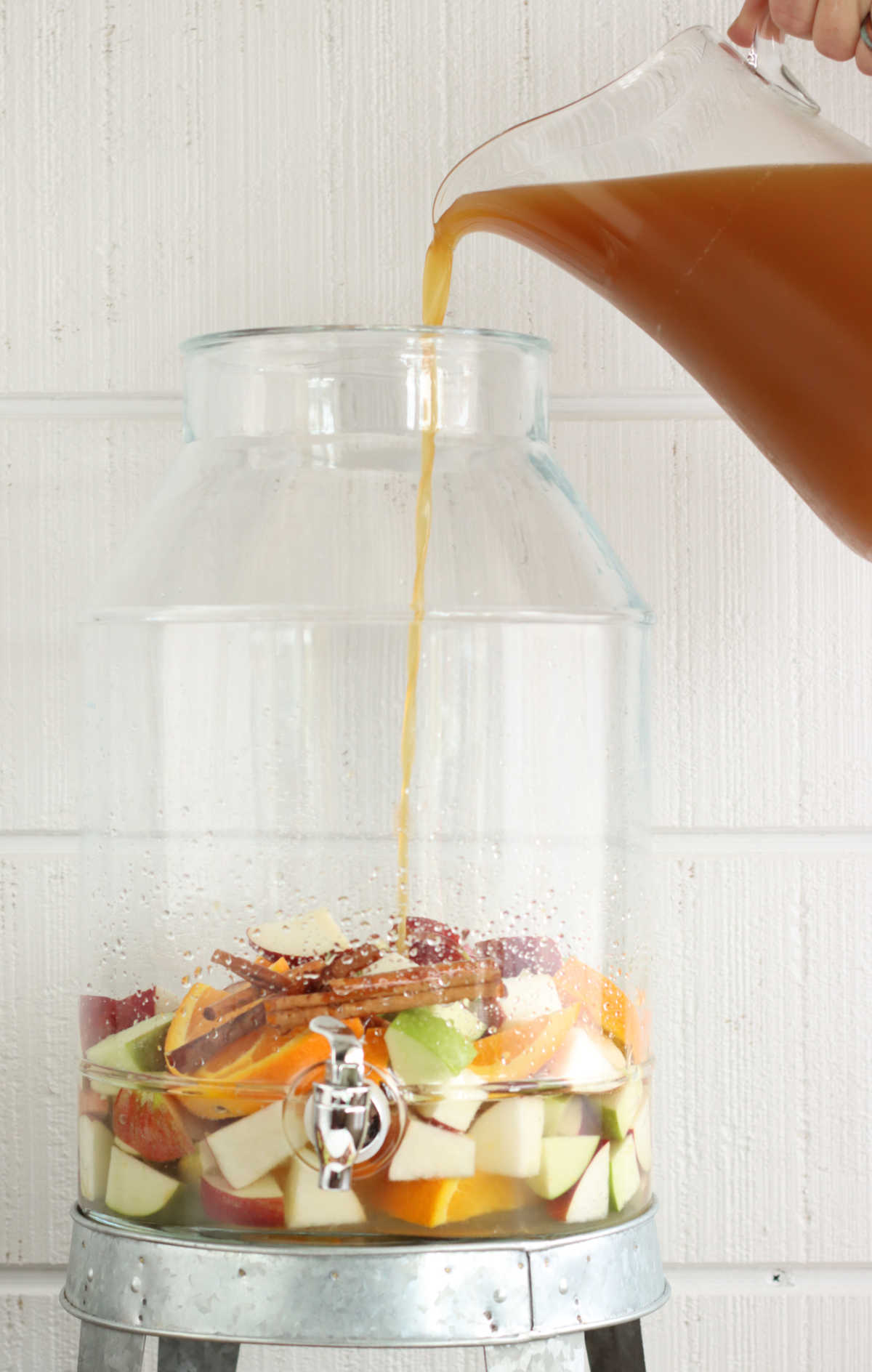 Pouring apple cider into glass drink dispenser filled with chunks of apples and oranges.