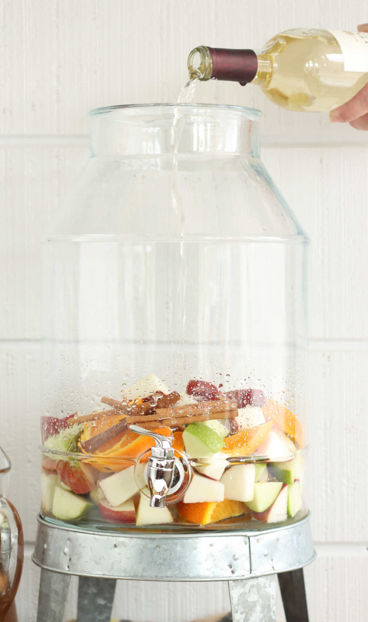 Pouring white wine into clear glass drink dispenser filled with apple and orange chunks.