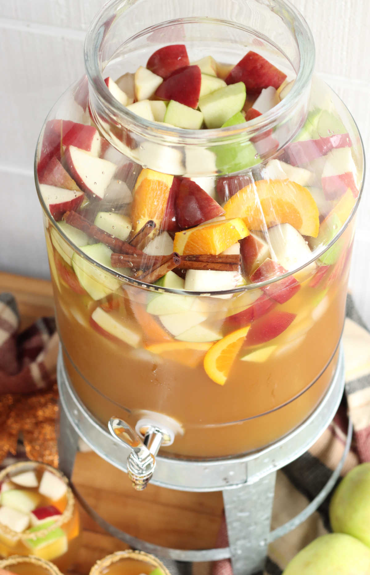Cider sangria in clear drink dispenser with chunks of apples and oranges, cinnamon sticks on galvanized stand.