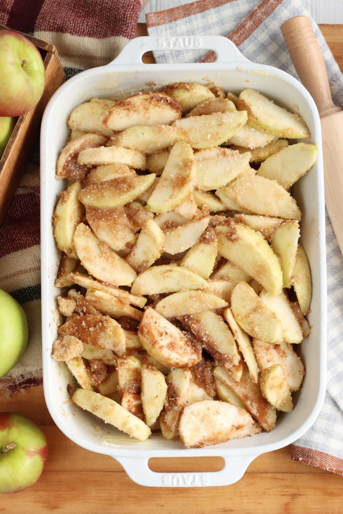 Apple slices, cinnamon sugar in white baking dish on wooden cutting board, fresh apples around, rolling pin.