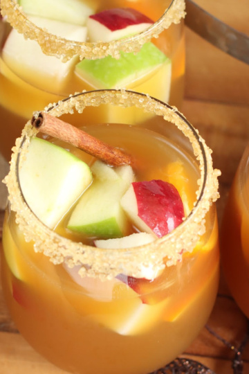 Apple sangria in stemless wine glasses, rims with sugar, apples, oranges, cinnamon sticks in glass.