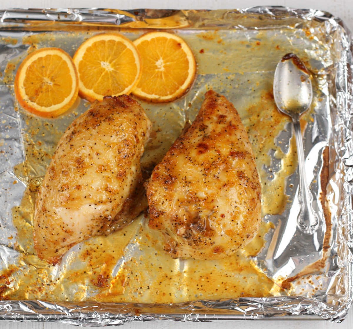Orange glazed chicken on a half sheet pan lined with aluminum foil and orange slices.