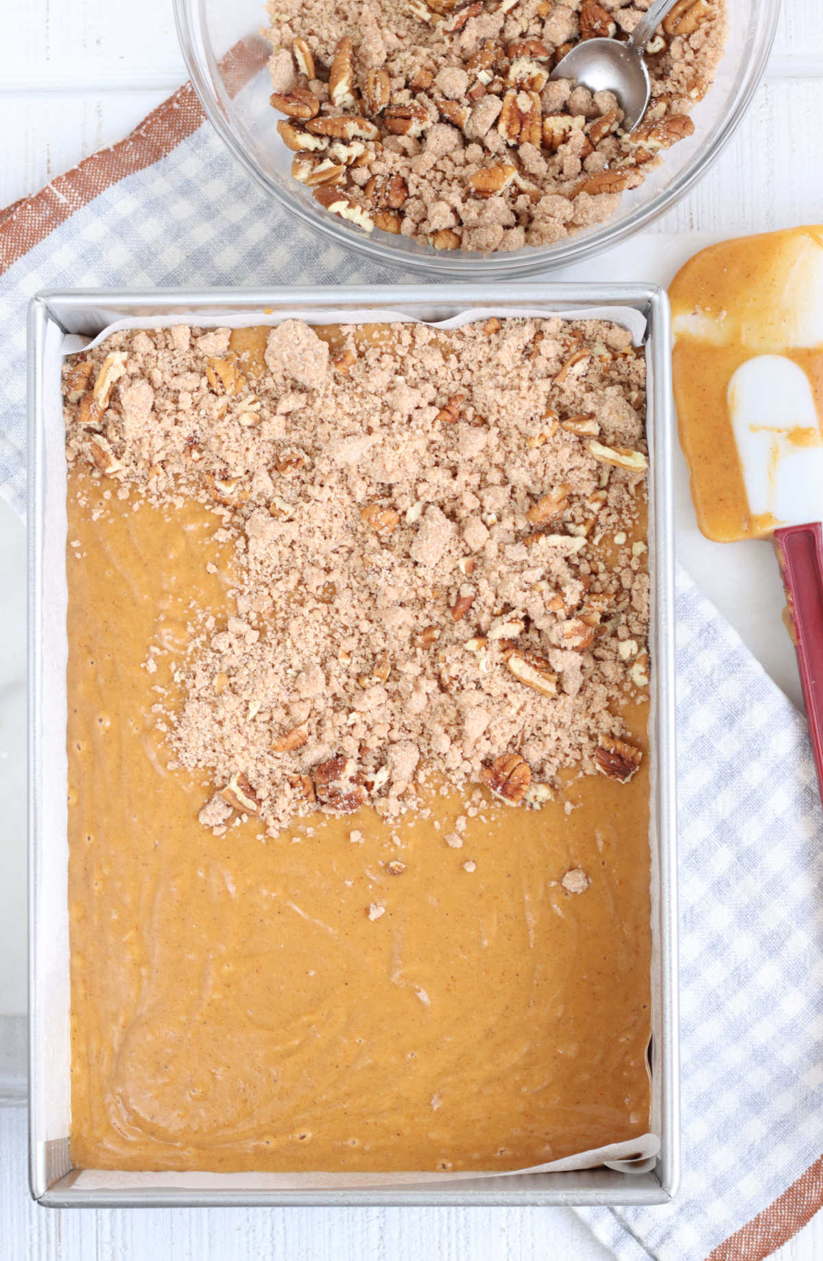 Pumpkin cake batter in rectangle metal baking pan topped with crumb and pecans, spatula to right.
