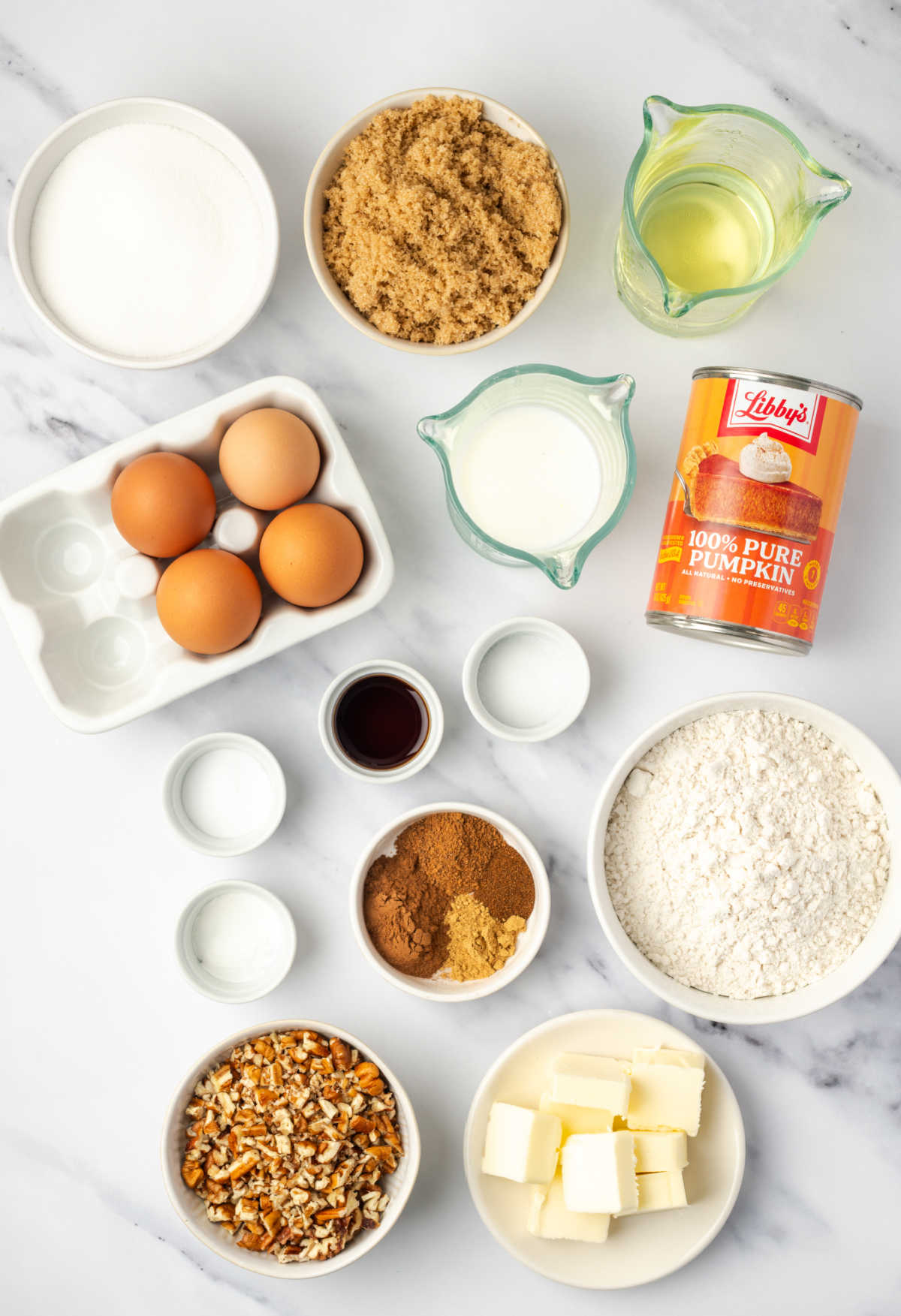 Ingredients in small white bowls on white marble for pumpkin cake.