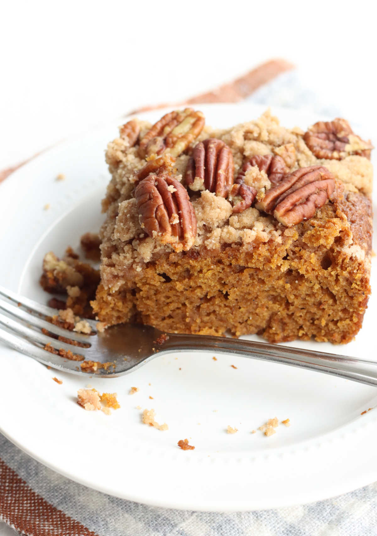 Square piece of pumpkin cake with crumb topping and pecans on small white plate with fork.