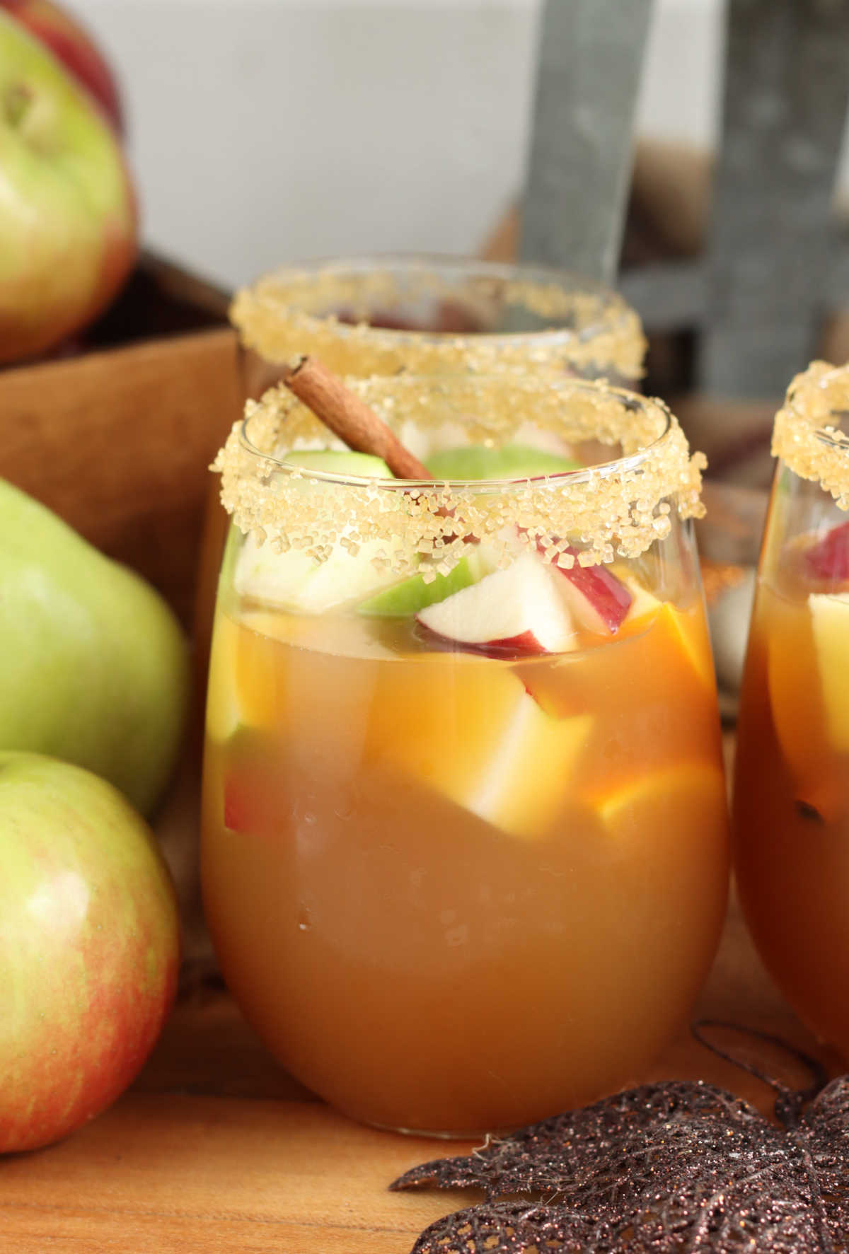 Stemless wine glass with apple sangria, chunks of apples and oranges, cinnamon stick on wooden cutting board, rimmed with raw sugar.