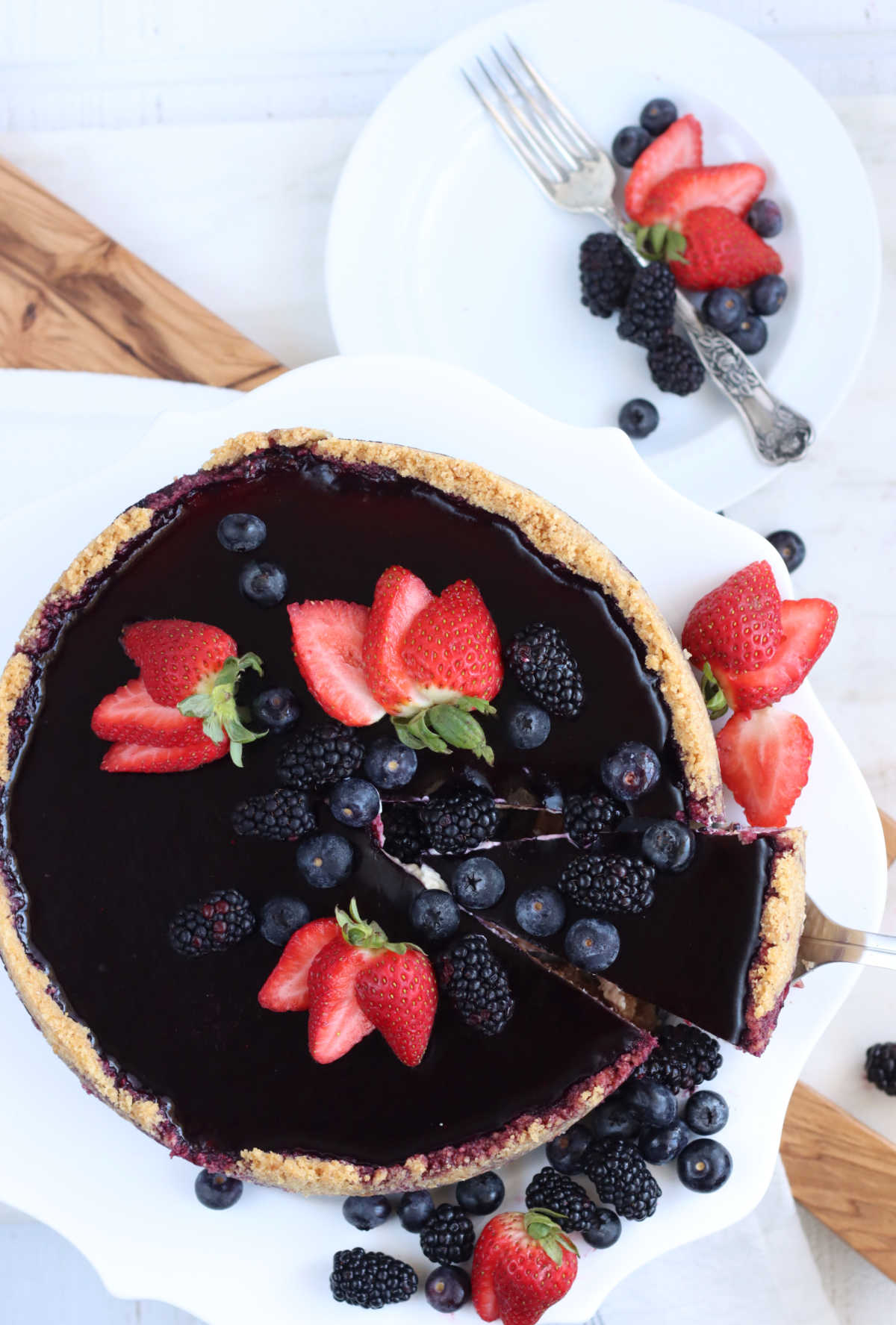 Cheesecake with berry topping on white footed cake dish, topped with strawberries, blackberries and blueberries, slice cut.