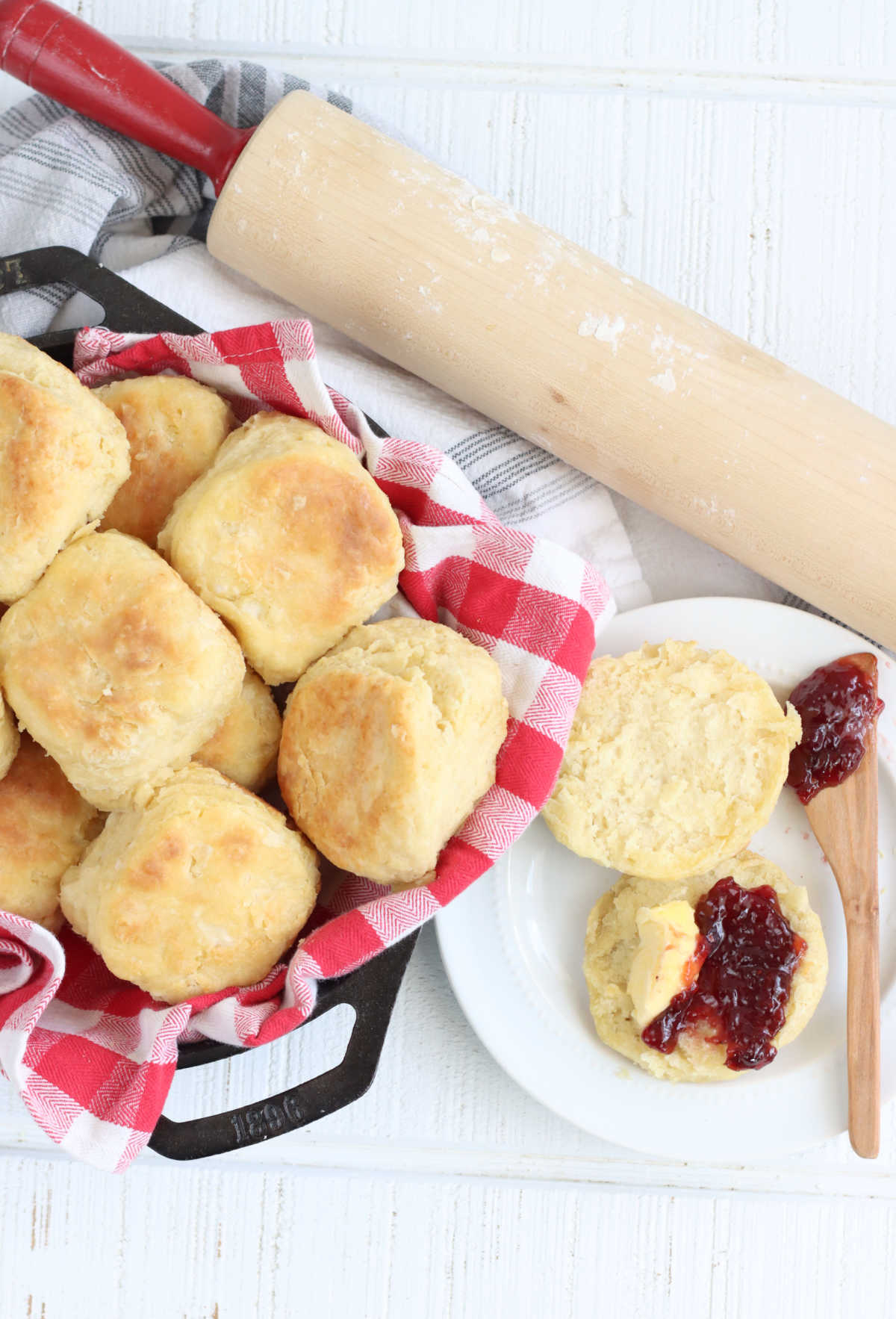 Biscuits in cast iron skillet with red checkered cloth, biscuit with jam on white plate to side.