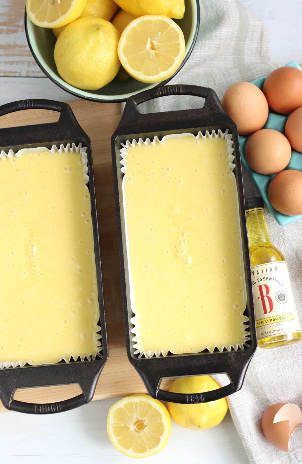 Lemon cake batter in two cast iron loaf pans on wooden cutting board, lemons around.