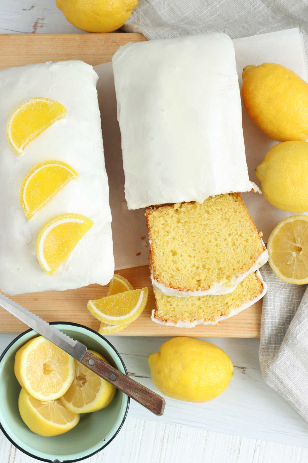 Two loaves of lemon cake, one partially sliced on wooden cutting board, fresh lemons.