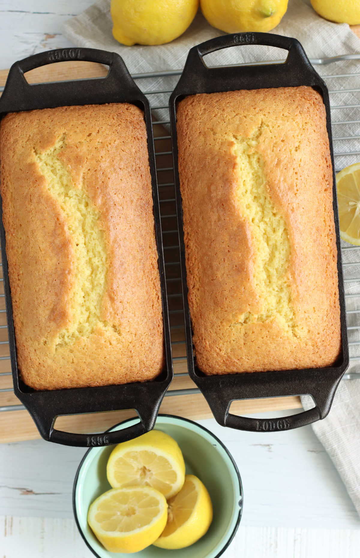 Two loaves of lemon cake in cast iron loaf pans on wooden cutting board, bowl of lemon slices.