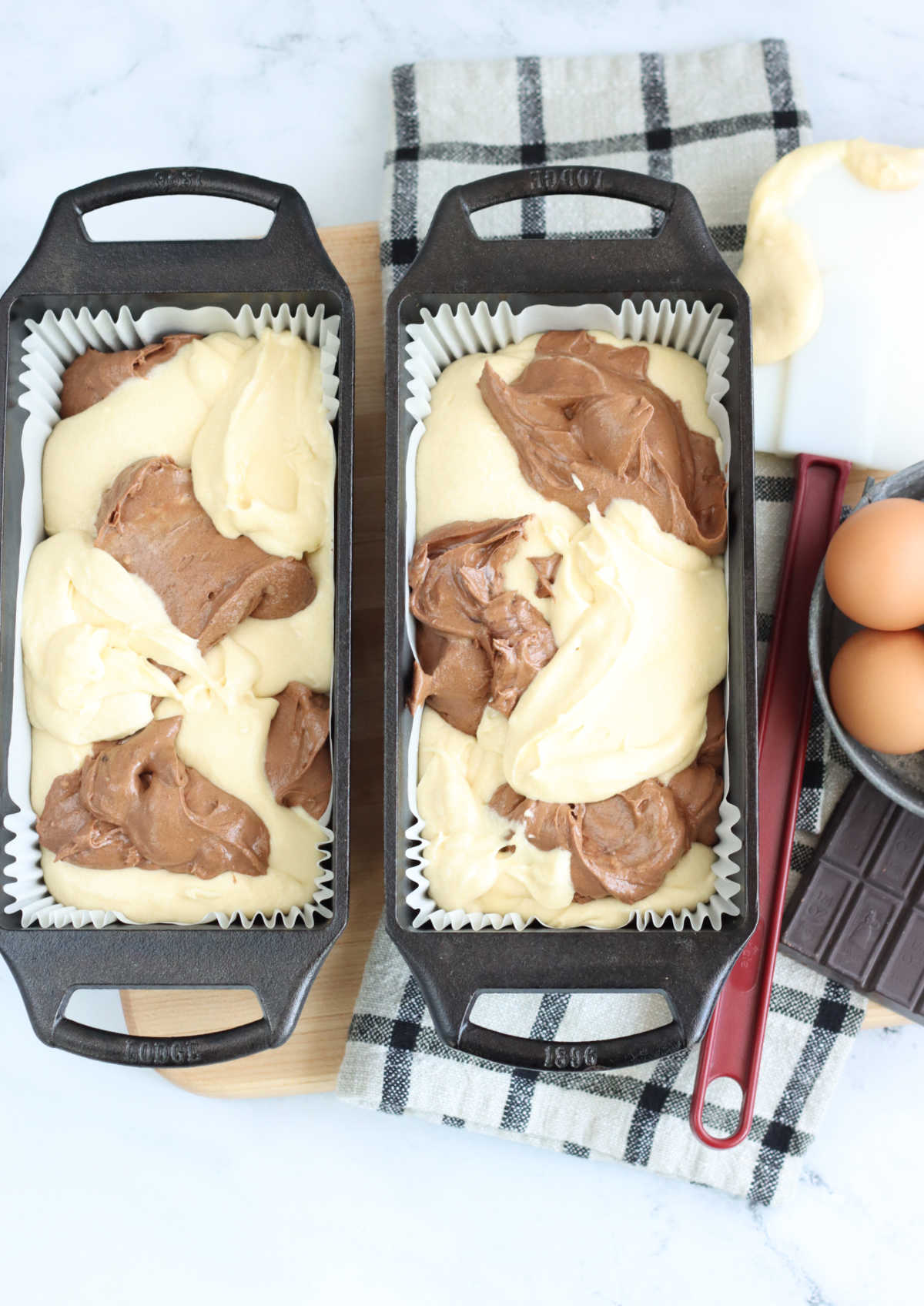 Two cast iron loaf pans with chocolate and vanilla cake batter scoops on wooden cutting board.