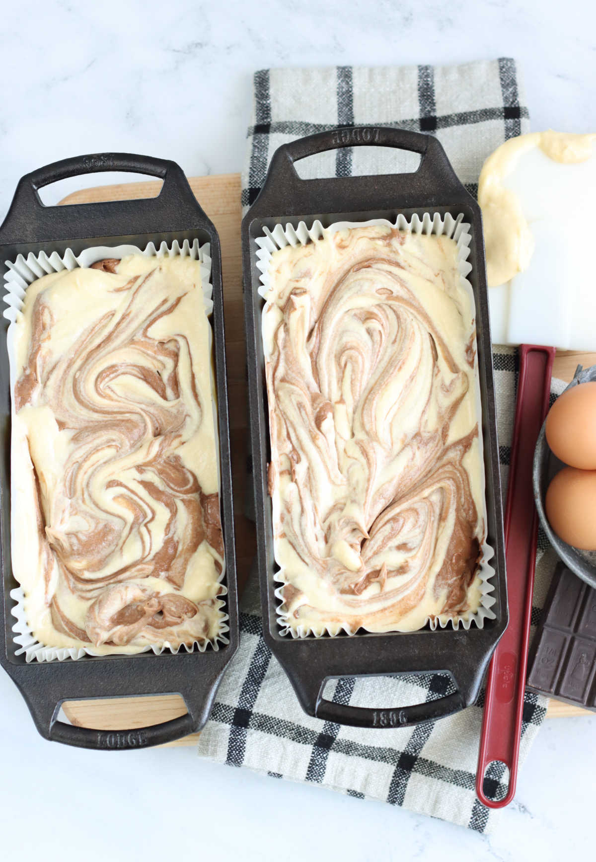 Chocolate and vanilla cake batter swirled in two cast iron loaf pans on wooden cutting board.