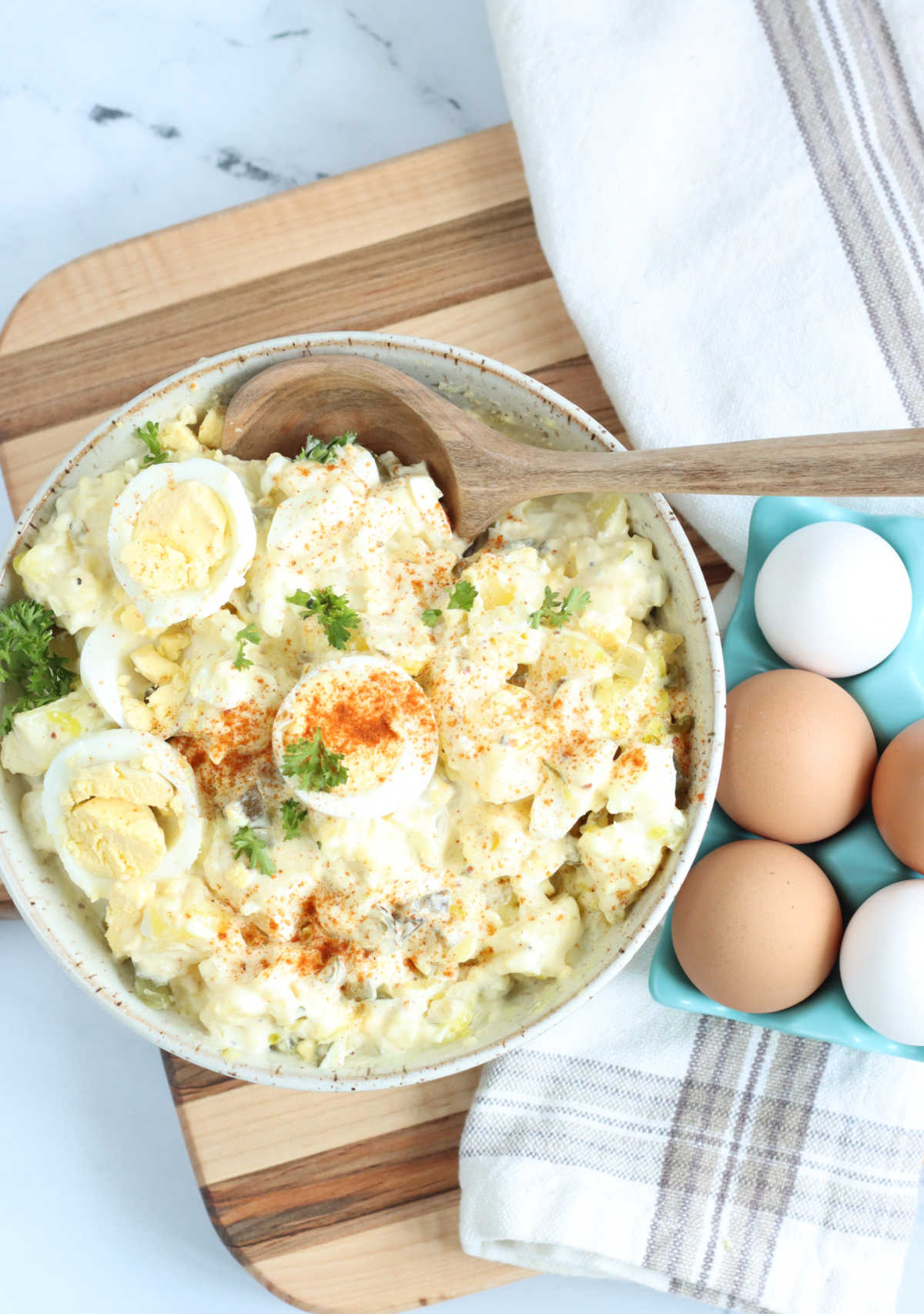 Bowl of potato salad with eggs on wooden cutting board.