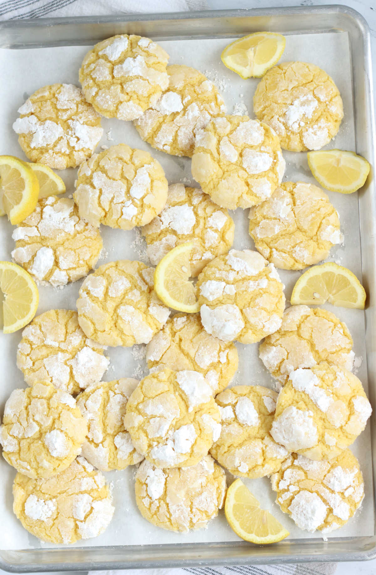 Lemon crinkle cookies on half sheet pan with lemon wedges.