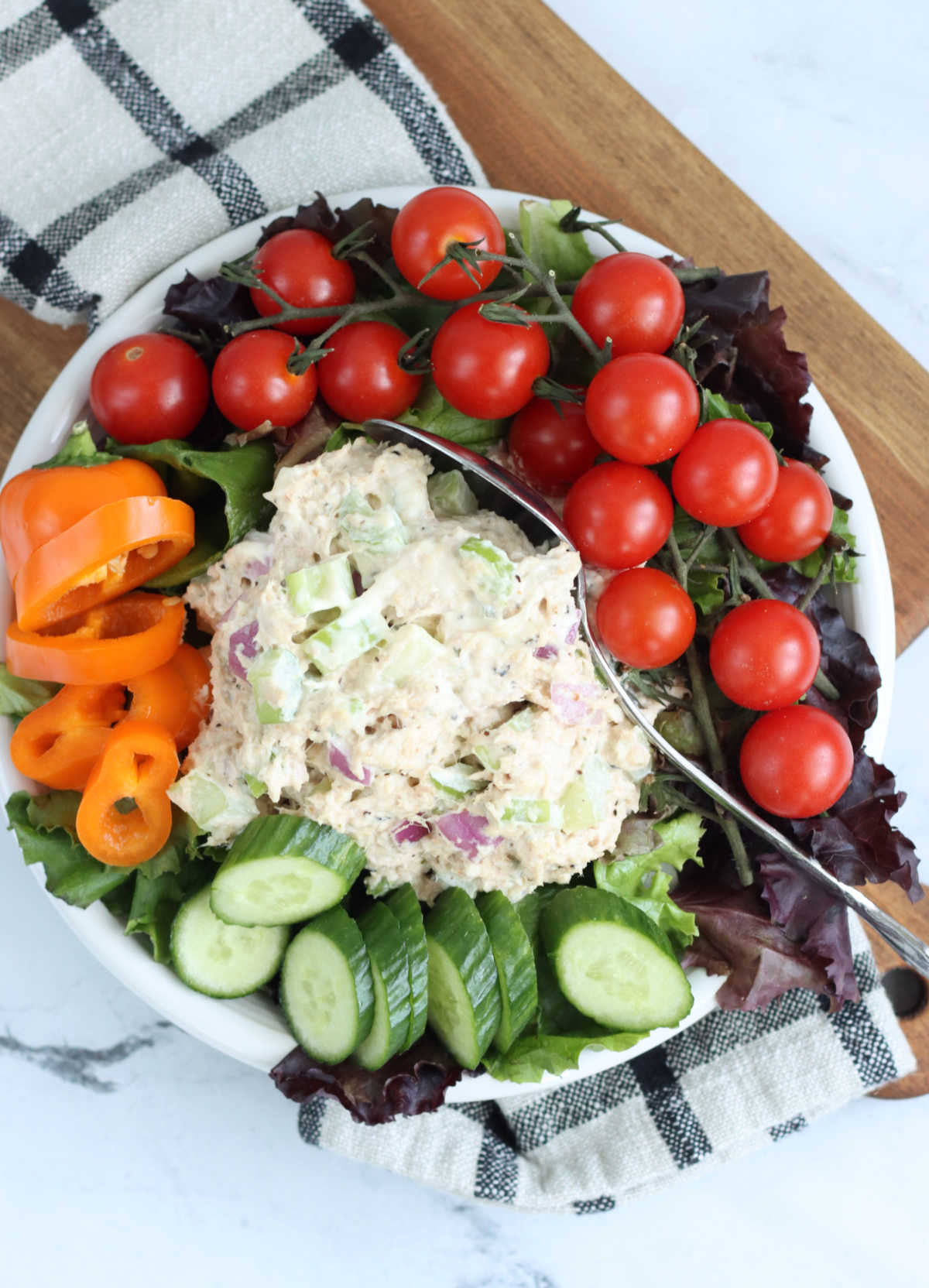 Scoop of tuna salad on small white plate of salad greens with cherry tomatoes, sliced cucumber and mini bell pepper.