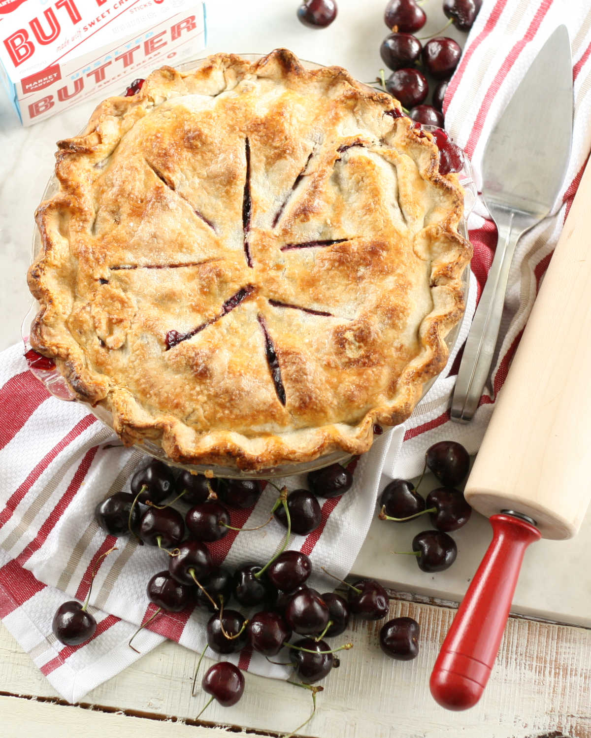 Cherry pie on white marble, loose fresh cherries around, red handled wooden rolling pin.